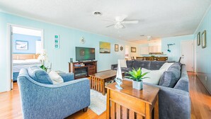 A spacious living room with a blue couch, a grey sofa, and a wooden coffee table. There is a television, a bookshelf, and a dining area visible in the background. - Landing Pad - Kill Devil Hills, Oceanside, 350 Yards to Beach, YR Flex Stays, 4 BR (Kill Devil Hills)