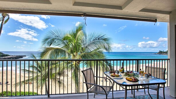 Un balcón con una mesa lista para comer, con vista a una playa con palmeras y el océano.