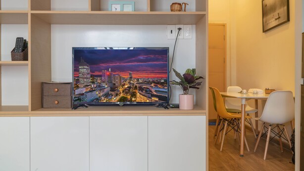 Ein modernes Wohnzimmer mit einem Flachbildfernseher auf einem Holzregal, einem Essbereich mit Tisch und Stühlen sowie einer Topfpflanze.