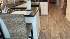 A modern kitchen with a central island, a dining area with wicker chairs, and a sign that reads "BEACH BEST ESCAPE ANYONE CAN HAVE". - Riverfront A-Frame w/ Dock, Kayaks, Putt-Putt & Shuffleboard (Oscoda Twp)