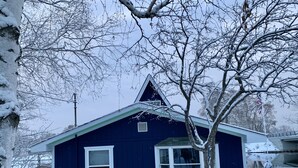 A dark blue house with a gabled roof and white trim, surrounded by snow-covered trees and a fence. - Riverfront A-Frame w/ Dock, Kayaks, Putt-Putt & Shuffleboard (Oscoda Twp)