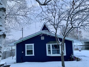 A dark blue house with a gabled roof and white trim, surrounded by snow-covered trees and a fence.