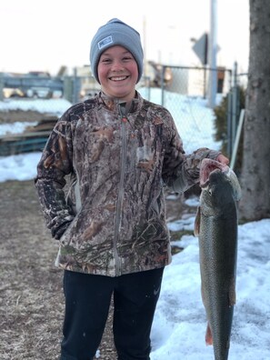 A person holding a fish, wearing a camouflage jacket and a beanie, standing outdoors with snow on the ground.