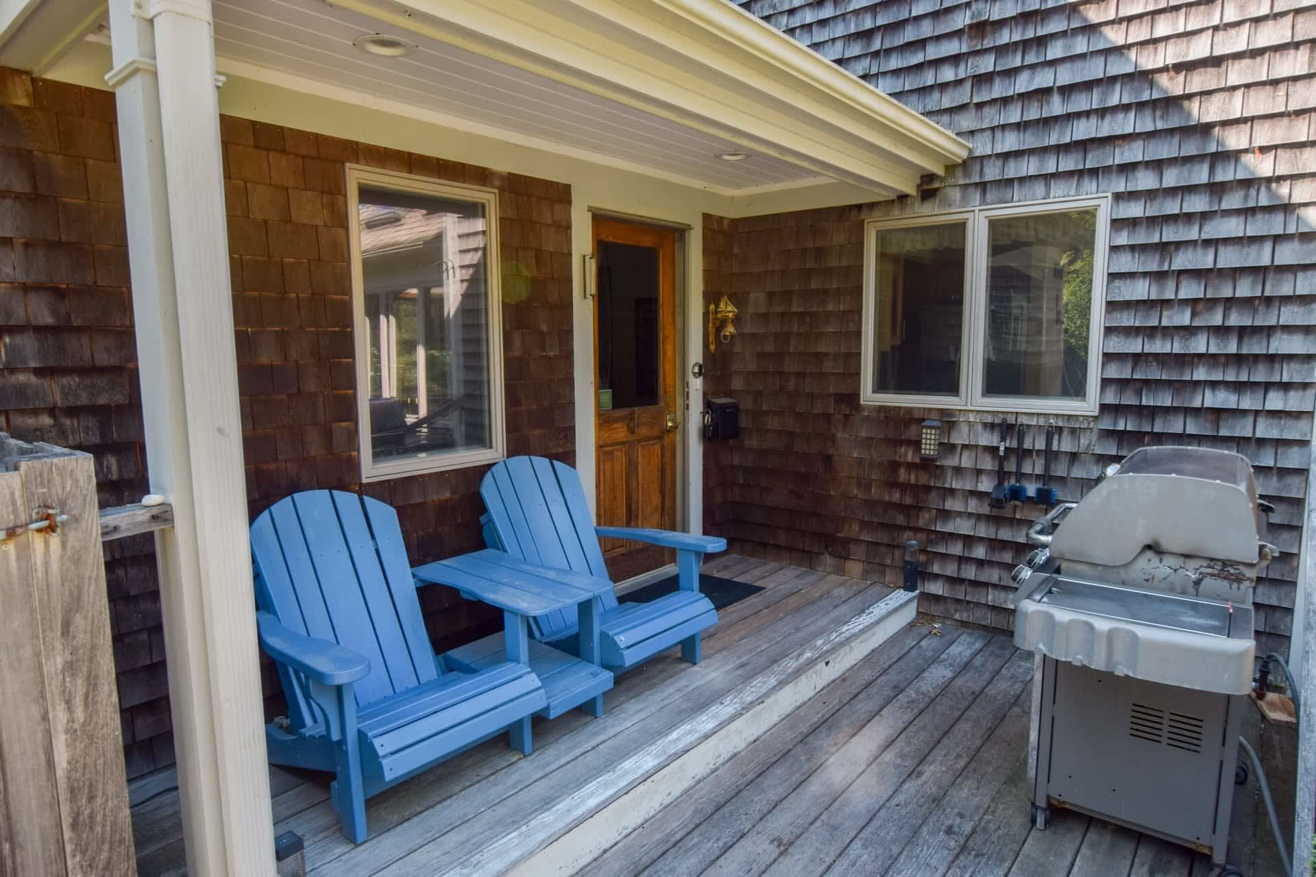 A covered porch with two Adirondack chairs, a grill, and a wooden door.