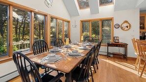 A dining area with a long wooden table set for six, surrounded by black chairs, large windows, and a skylight.