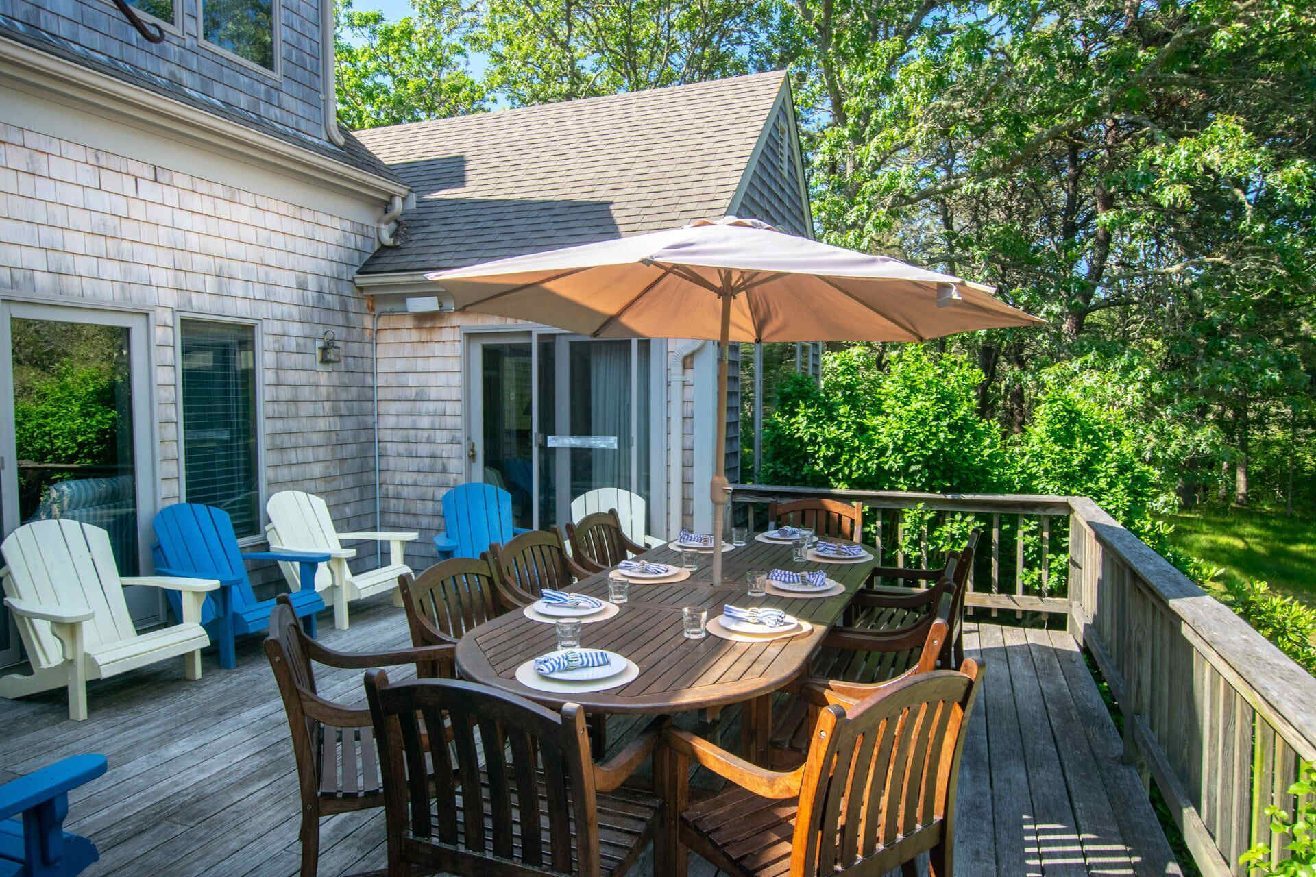 A wooden deck with a table set for six, surrounded by chairs, and an umbrella providing shade.