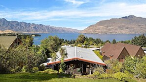 Una cabaña junto al lago con techo de metal, rodeada de vegetación exuberante y montañas al fondo.