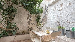 Un pequeño espacio exterior para comer con una mesa y sillas blancas, rodeado de plantas en macetas y una pared con textura.