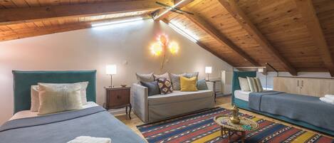 A cozy attic bedroom with a sloped ceiling, wooden beams, a large bed with a blue headboard, a sofa, and a colorful rug.