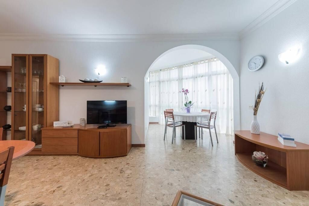 A spacious living area with a TV, wooden cabinets, a dining table, and a large window with natural light.