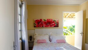 A bedroom with a bed, a nightstand, a window with curtains, and a painting of red flowers on the wall. - Pousada Luzes de Geribá (Búzios)