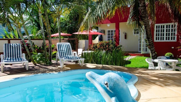 A swimming pool with lounge chairs and umbrellas, surrounded by greenery and a red building. - Pousada Luzes de Geribá (Búzios)