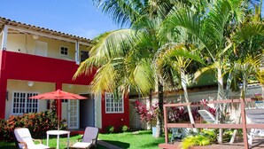 A red and yellow building with a patio area featuring two white chairs, a red umbrella, and a small table. - Pousada Luzes de Geribá (Búzios)