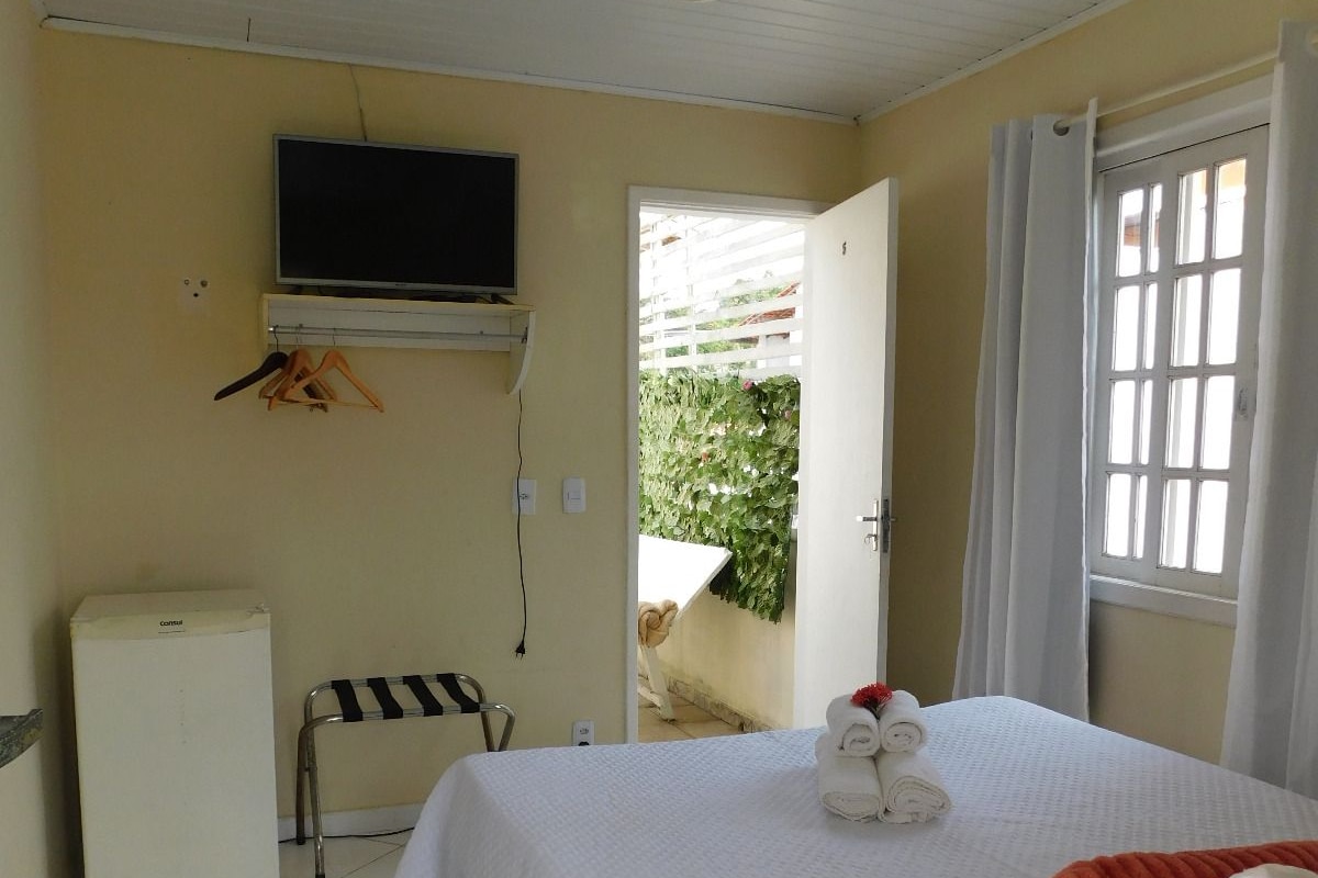 A bedroom with a bed, a television mounted on the wall, a small refrigerator, and a window with curtains.