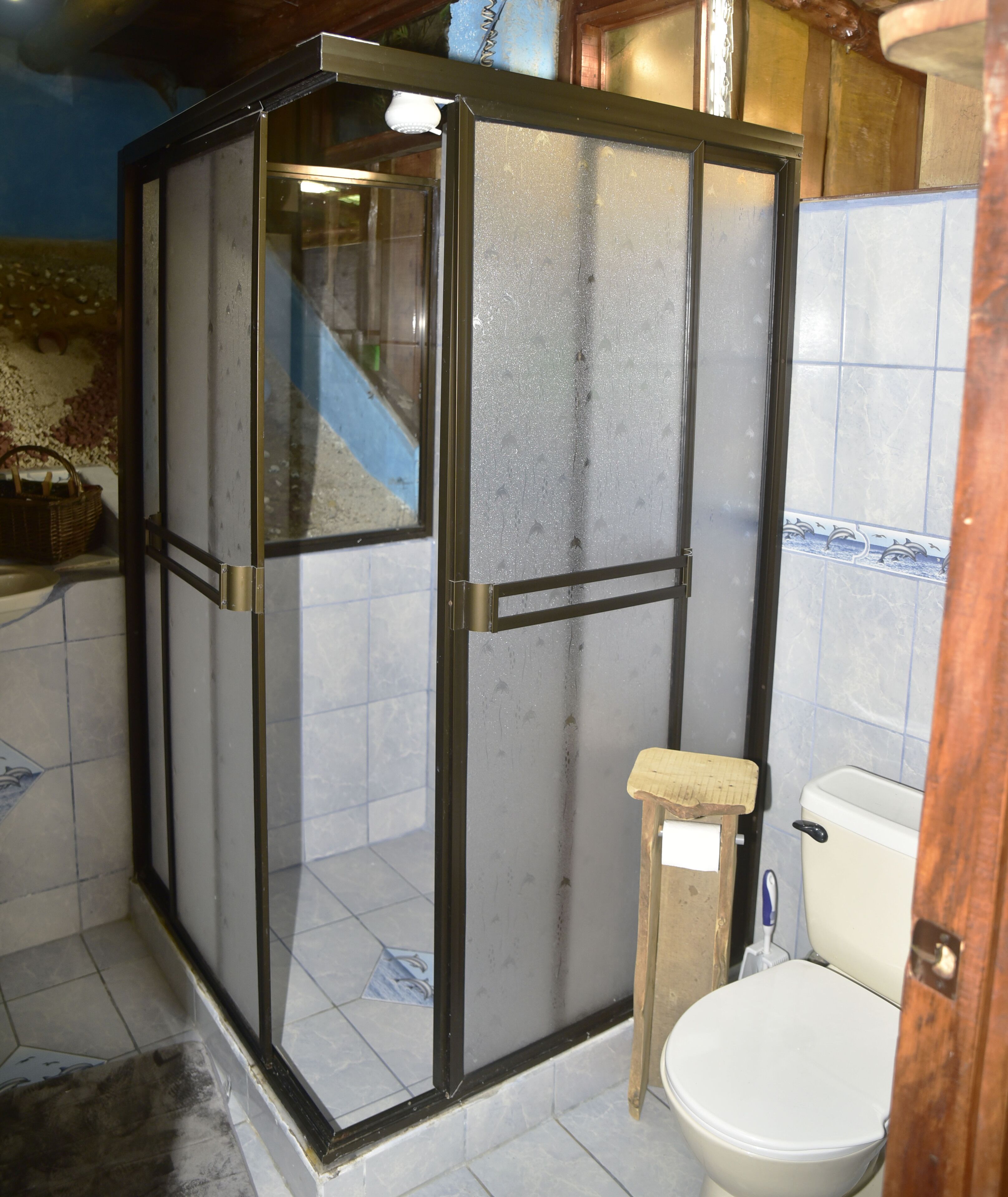 Un bany amb una dutxera de vidre, un vàter i un petit tamboret de fusta.