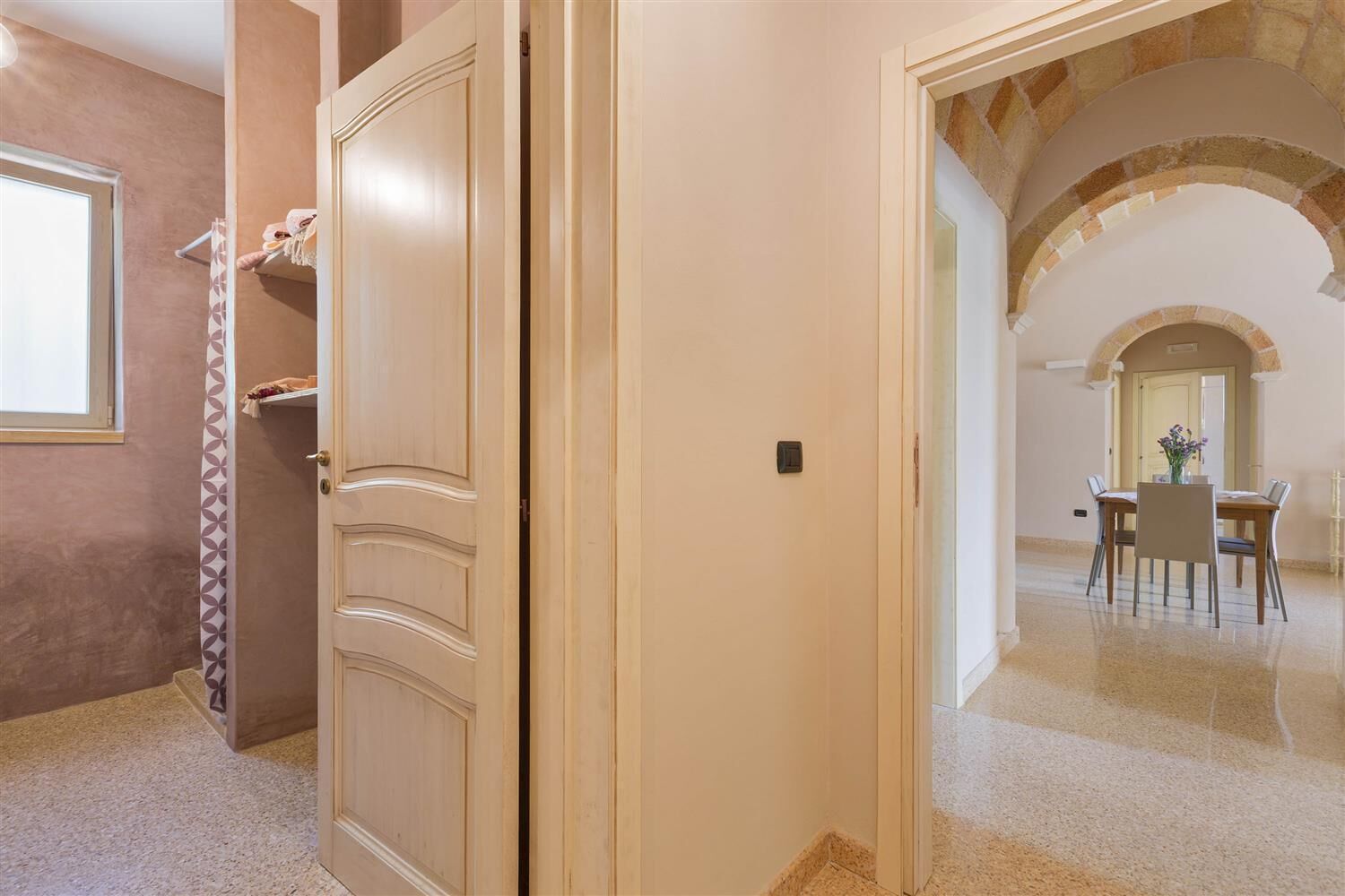 Un pasillo con una puerta cerrada, un cortinero estampado y una zona de comedor con mesa y sillas.
