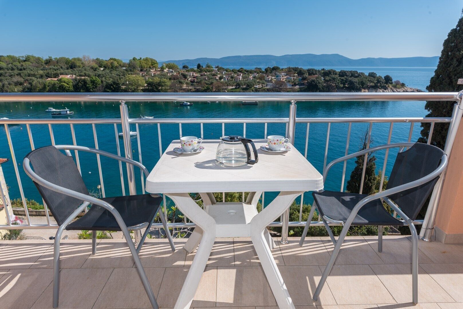 A balcony with a table and chairs overlooking a body of water with boats and a distant shoreline.