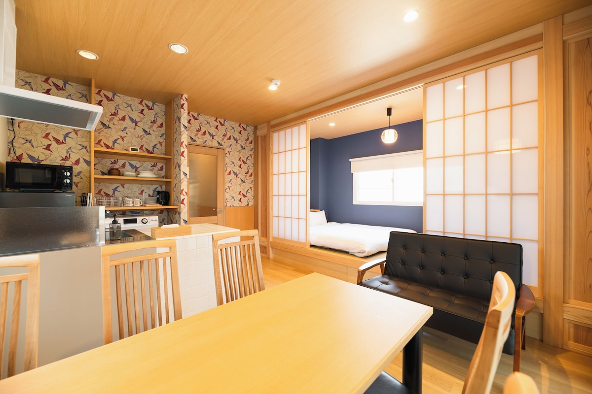 A compact room with a kitchenette, a dining table, a sofa, and a bed with a view of the outside.