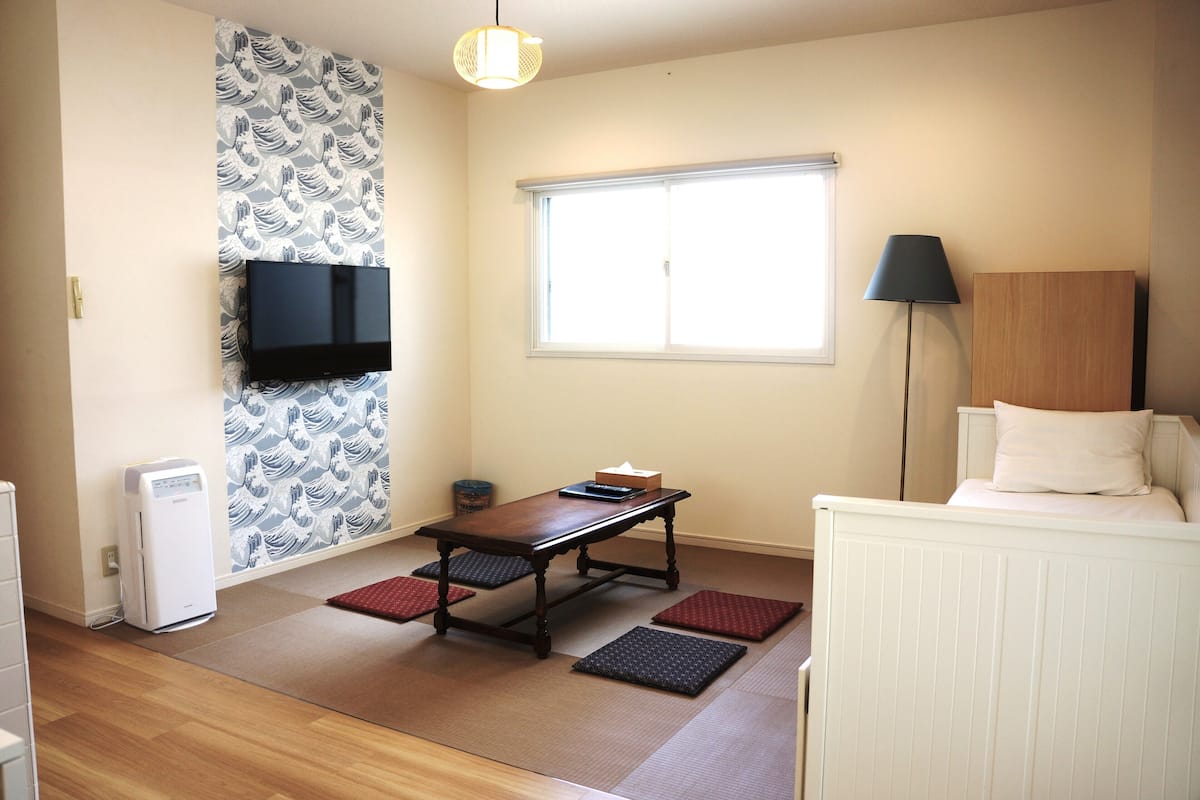 Ein Zimmer mit Tatami-Matten, einem niedrigen Tisch, einem an der Wand montierten Fernseher, einem Fenster mit Jalousien und einer Nachttischlampe.