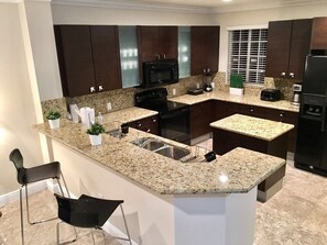 A modern kitchen with granite countertops, dark wooden cabinets, a built-in microwave, a stove, and a sink. There are two bar stools and a window with blinds. - 4BR Townhouse 906 by Ideal Experience VR (Fort Lauderdale)