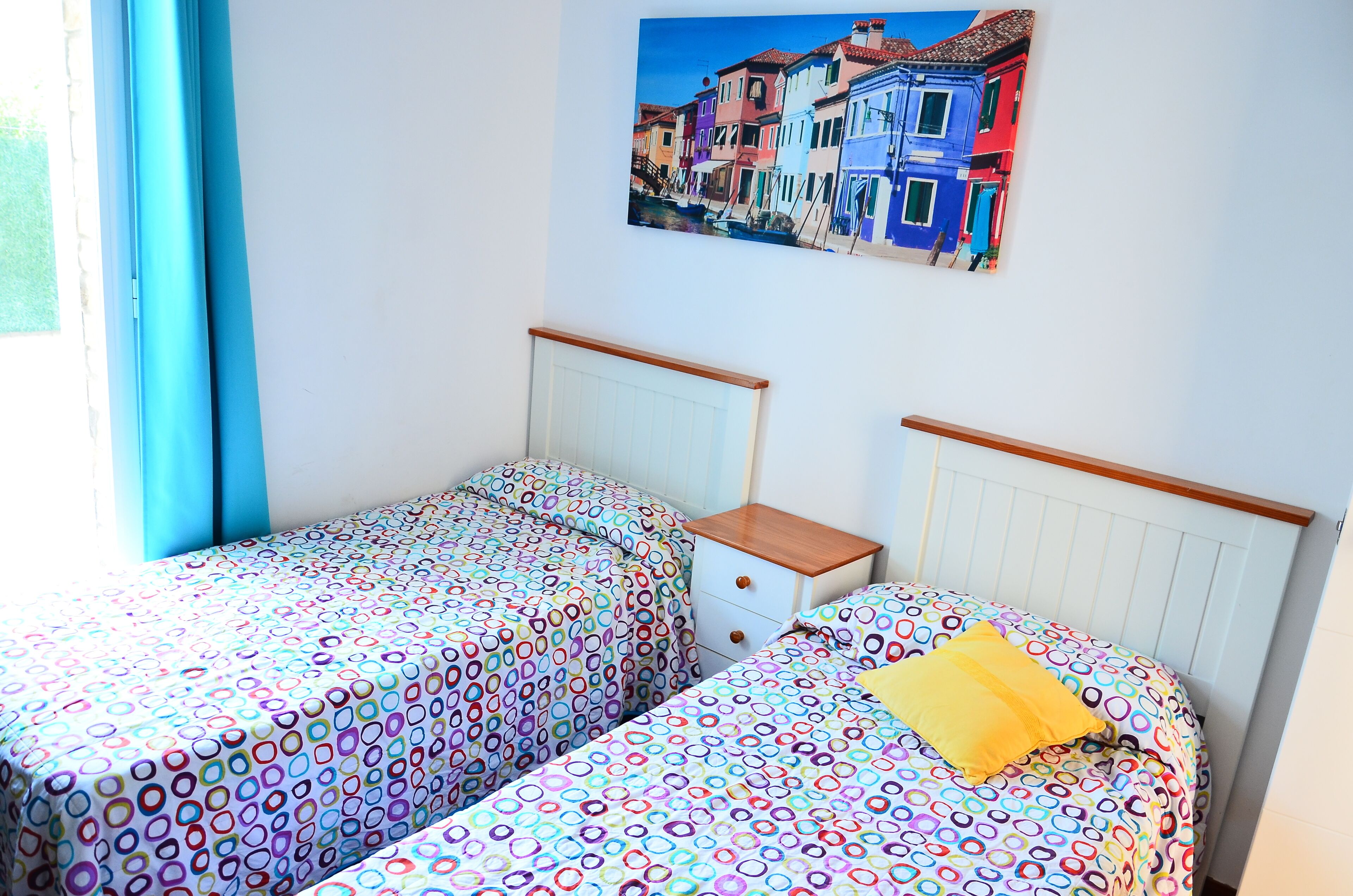 A room with two beds, a nightstand, and a wall-mounted painting of colorful buildings.