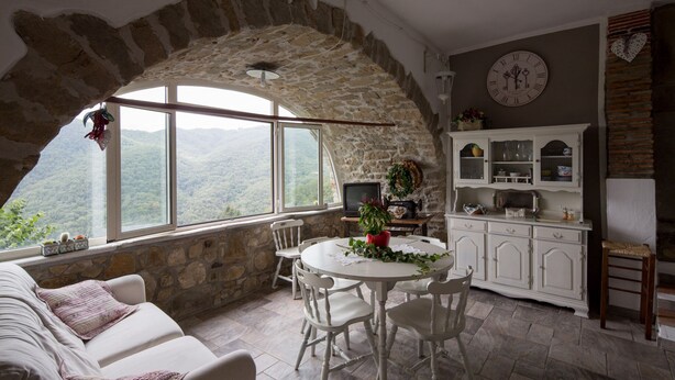 A rustic stone-walled room with a dining table and chairs, a white sofa, and a kitchen area with cabinets and a clock. - Stone Rustico with Jacuzzi, Infrared Sauna Escluiva   (Boveglio)