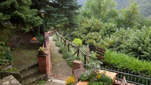 A rustic outdoor seating area with a brick wall, wooden table, and chairs, surrounded by lush greenery and a stone pathway. - Stone Rustico with Jacuzzi, Infrared Sauna Escluiva   (Boveglio)