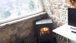 A stone-walled room with a wood-burning stove, a window with a view of a forest, and a table with a chair. - Stone Rustico with Jacuzzi, Infrared Sauna Escluiva   (Boveglio)