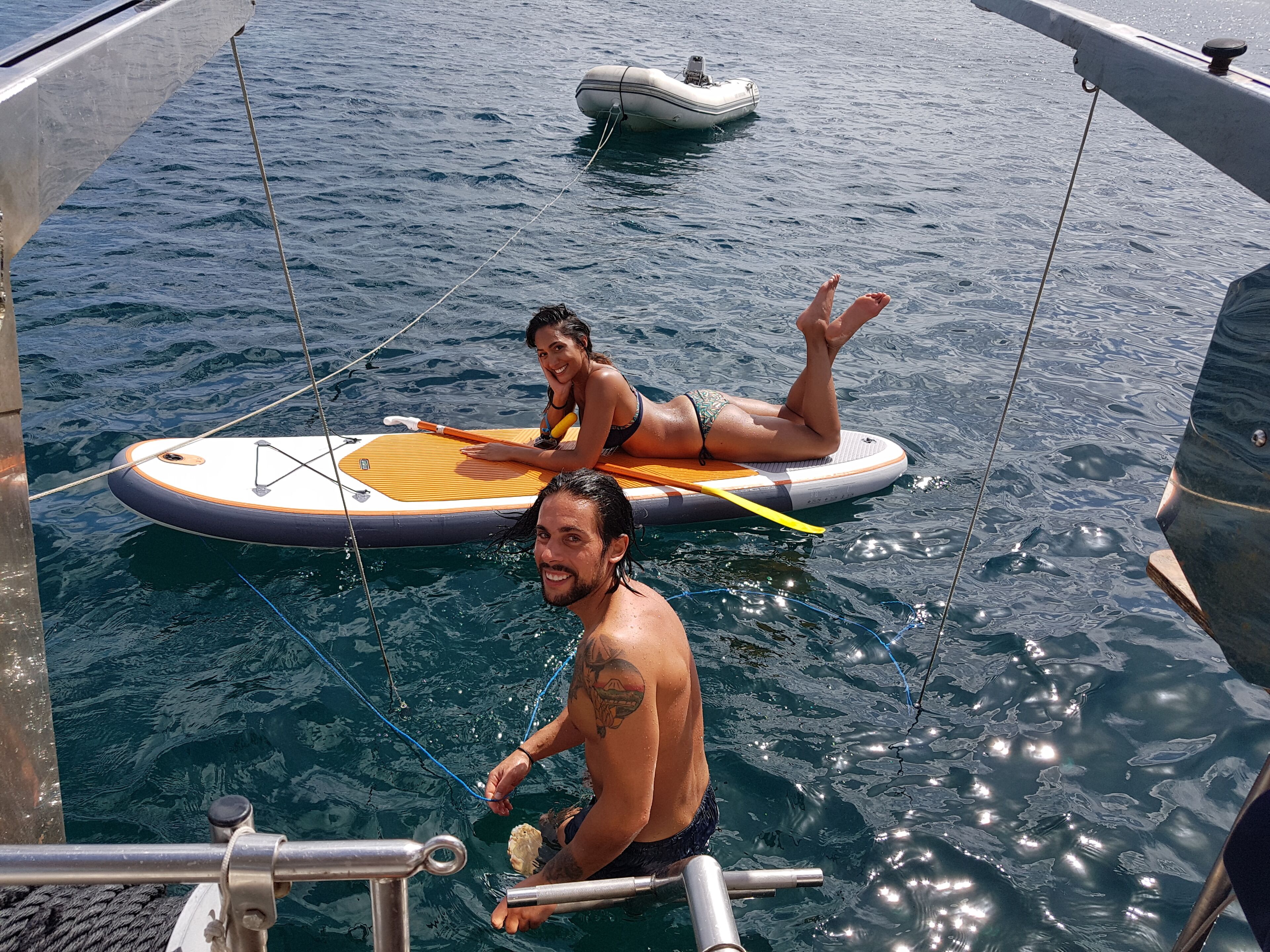 A boat with two people on a surfboard, a smaller inflatable boat in the background, and a metal railing.