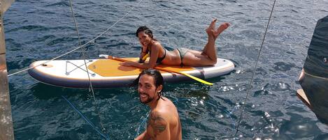 A boat with two people on a surfboard, a smaller inflatable boat in the background, and a metal railing.