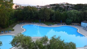 A large outdoor swimming pool with multiple lounge chairs and umbrellas, surrounded by trees and residential buildings in the background.