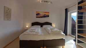 A neatly made bed with pillows and a folded blanket, a bedside table with a lamp, a painting of a mountain landscape, and a window with a view of the outdoors. - The Steinbock Lodge (Zell am See)