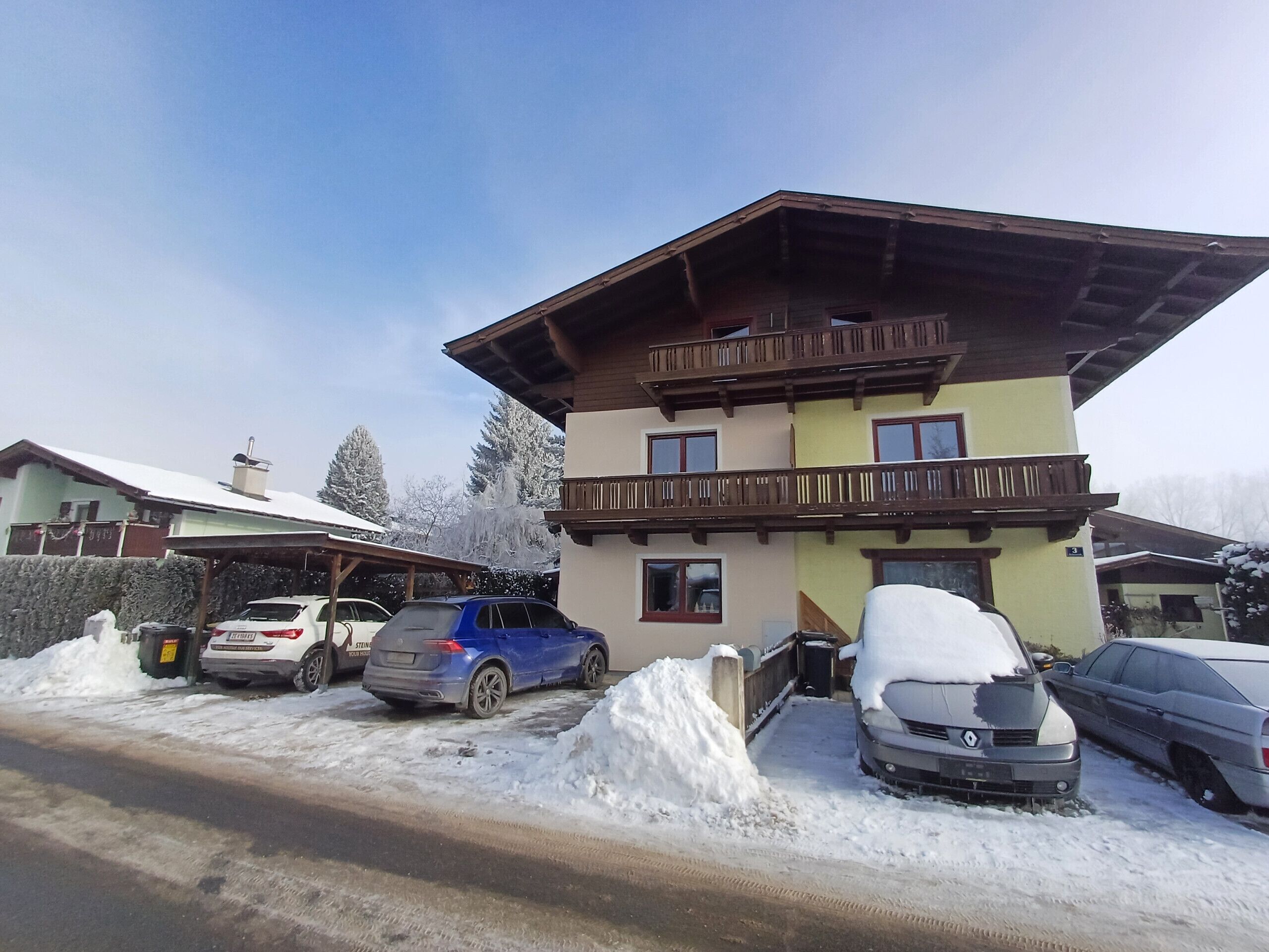 A multi-story building with a sloped roof and wooden balconies. There are several cars parked in front, some covered in snow.