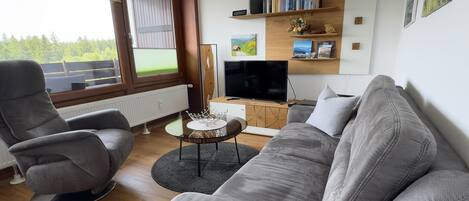 A modern living room with a grey sofa, two armchairs, a coffee table, and a TV. There are bookshelves with books and decorative items.
