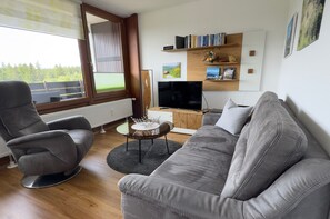 Ein modernes Wohnzimmer mit einem grauen Sofa, zwei Sesseln, einem Couchtisch und einem Fernseher. Bücherregale mit Büchern und Dekorationselementen sind ebenfalls vorhanden.