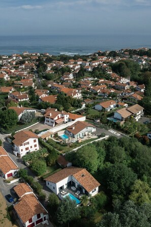 An aerial view of a coastal residential area with houses featuring red-tiled roofs and white walls, surrounded by greenery and a clear view of the sea.