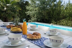 A breakfast setup with croissants, bananas, pineapple, and orange juice by a poolside with a view of trees.