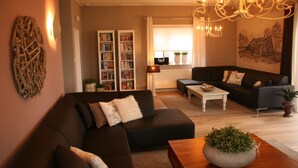 Ein modernes Wohnzimmer mit einem dunkelbraunen Ecksofa, einem weißen Couchtisch und einer Chandelieber. Bücherregale voller Bücher und ein Gemälde an der Wand sind ebenfalls vorhanden.
