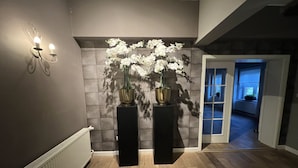 Ein Korridor mit einem Heizkörper, zwei schwarzen Podesten mit weißen Orchideen und einer wandmontierten Leuchte.
