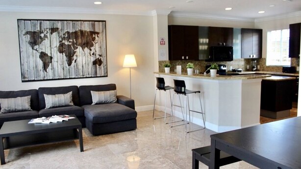 A modern living area with a sofa set, a coffee table, and a dining table. There is a kitchen area with dark cabinets and a microwave. A world map artwork is on the wall, and a lamp is on a side table. - 4BR Townhouse 320 by Ideal Experience VR (Fort Lauderdale)