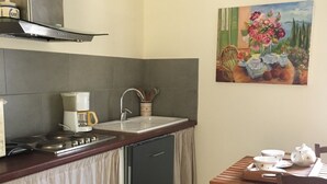 A compact kitchen with a gas stove, a coffee maker, a sink, and a small refrigerator. There is a dining table with chairs and a painting on the wall.