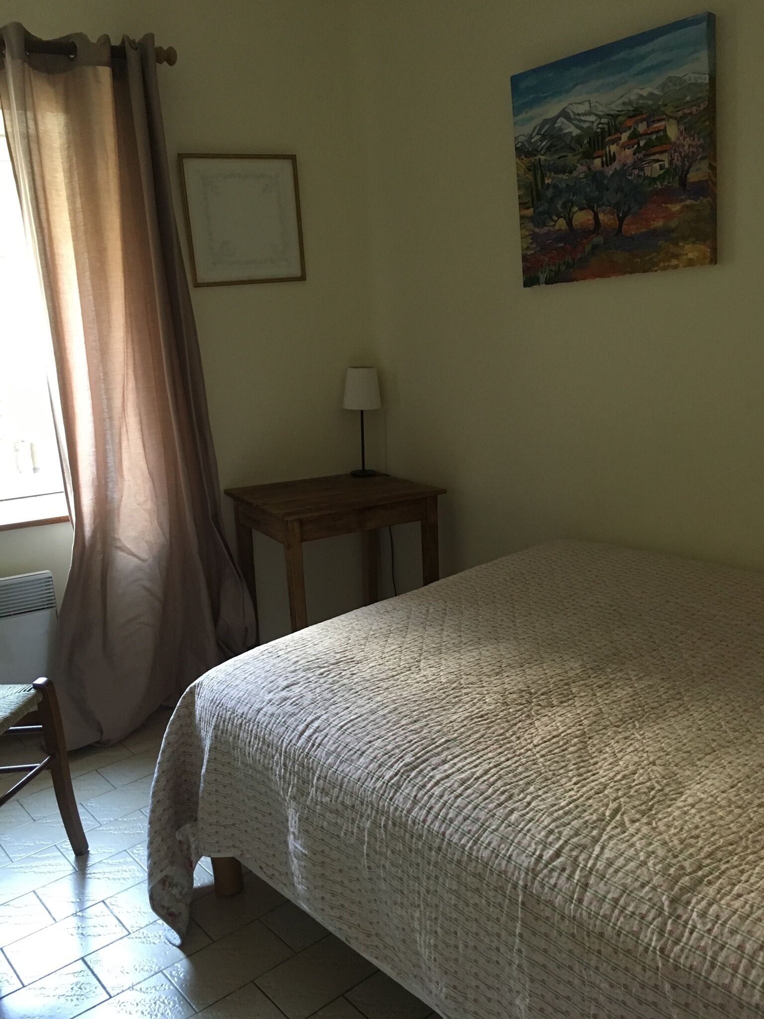 A bedroom with a bed, a nightstand, a lamp, a painting on the wall, and a window with curtains.