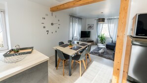 Moderni keittiö, jossa on puupalkkinen katto, ruokailutila pöydän ja tuolien kera, televisio sekä oleskelutila, jossa on sohva ja viherkasvi.