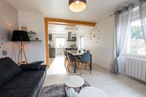 A modern living room with a sofa, a dining table with chairs, a lamp, and a view of the kitchen through an open doorway.