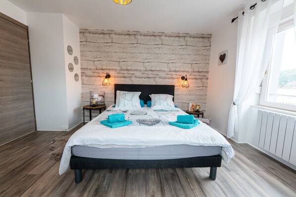 A modern bedroom with a wooden floor, a large bed with white bedding, two bedside tables with lamps, and a brick-patterned wall.