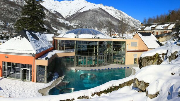A hotel with a glass-enclosed pool, snow-covered surroundings, and mountains in the background.