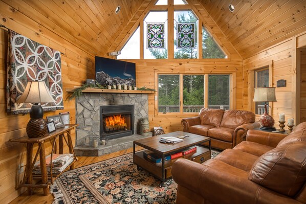 A cozy log cabin interior with a stone fireplace, leather furniture, and large windows offering a view of the forest.