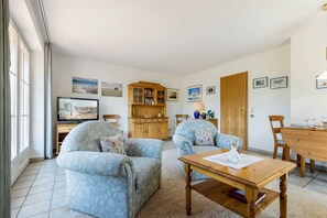 A living room with two floral-patterned armchairs, a wooden coffee table, a TV, a bookshelf, and a dining table with chairs.