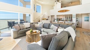 A spacious living area with a large gray sectional sofa, a round wooden coffee table, and a view of the ocean.