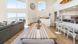 A spacious living area with a large window overlooking the beach, a wooden dining table, and a kitchen with white cabinets.